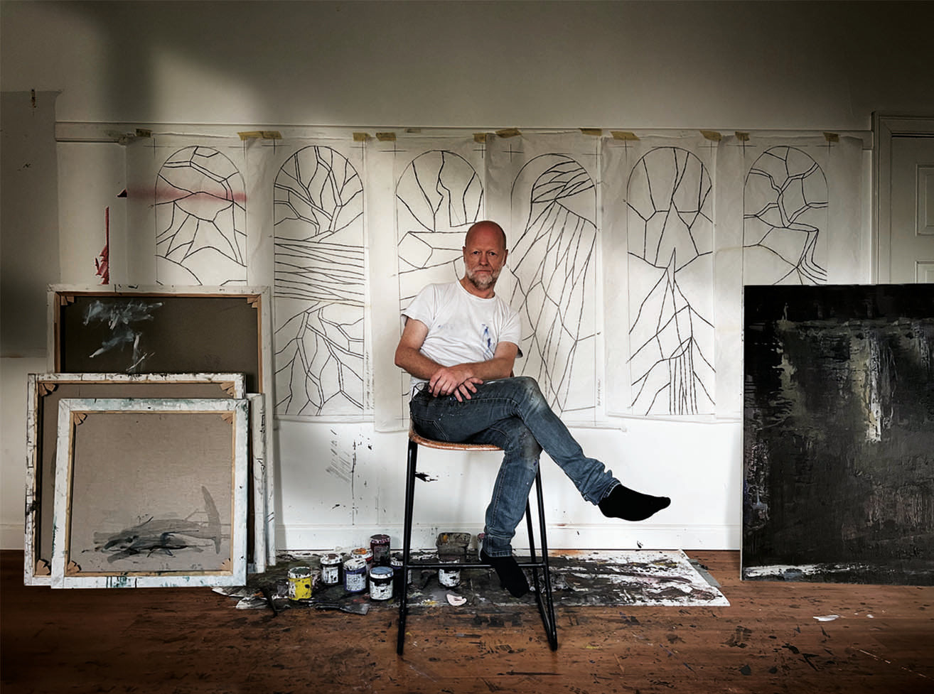 Kunstner Peter Ejlerskov sidder på taburet i sit atelier foran skitser og malerier